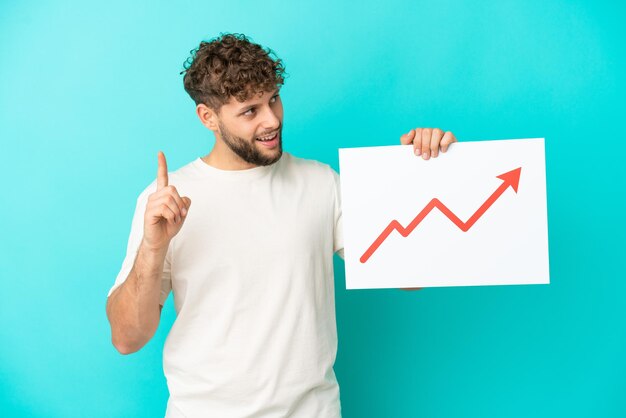 Young handsome caucasian man isolated on blue background holding a sign with a growing statistics arrow symbol and thinking