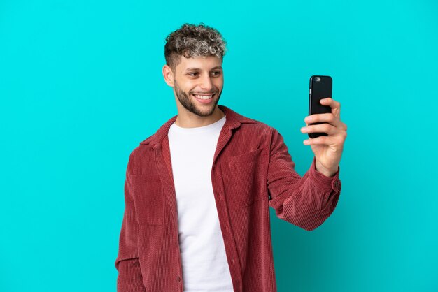 Young handsome caucasian man isolated on blue background making a selfie with mobile phone