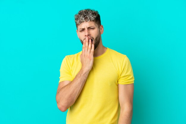 Young handsome caucasian man isolated on blue background yawning and covering wide open mouth with hand