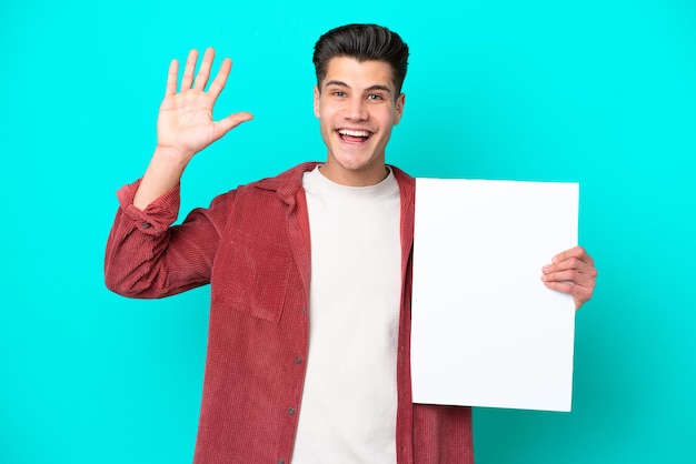 Young handsome caucasian man isolated on blue bakcground holding an empty placard and saluting