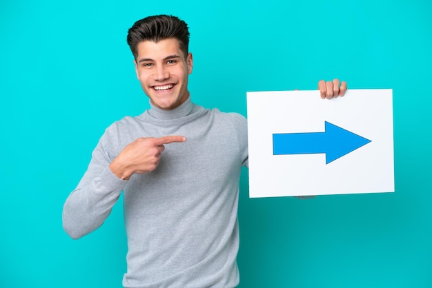 Young handsome caucasian man isolated on blue bakcground holding a placard with arrow symbol and pointing it