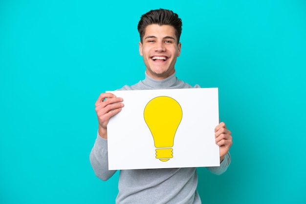 Young handsome caucasian man isolated on blue bakcground holding a placard with bulb icon with happy expression