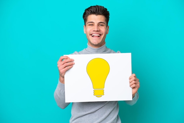 Giovane uomo caucasico bello isolato su bakcground blu che tiene un cartello con l'icona della lampadina con espressione felice