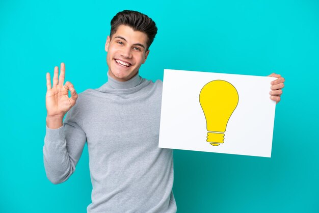Young handsome caucasian man isolated on blue bakcground holding a placard with bulb icon with ok sign