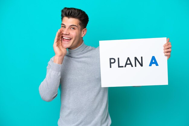 Young handsome caucasian man isolated on blue bakcground holding a placard with the message PLAN A and shouting