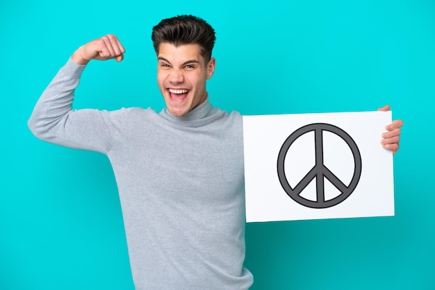 Young handsome caucasian man isolated on blue bakcground holding a placard with peace symbol and doing strong gesture