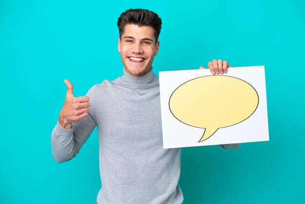 Young handsome caucasian man isolated on blue bakcground holding a placard with speech bubble icon with thumb up