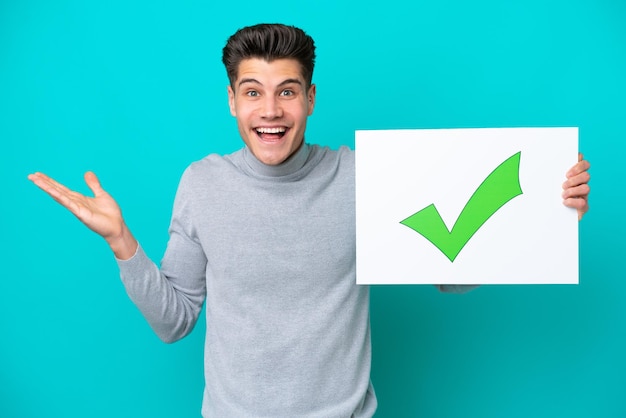 Young handsome caucasian man isolated on blue bakcground\
holding a placard with text green check mark icon with surprised\
expression