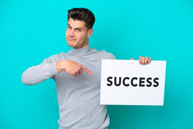 Young handsome caucasian man isolated on blue bakcground holding a placard with text SUCCESS and pointing it