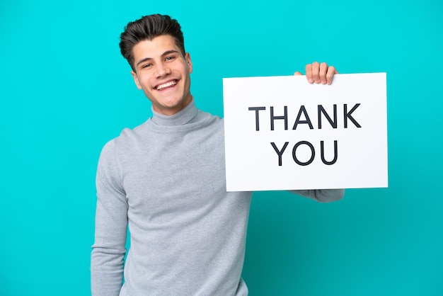 Young handsome caucasian man isolated on blue bakcground holding a placard with text thank you with happy expression