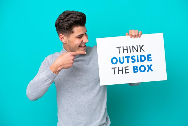 파란색 bakcground에 격리된 젊고 잘생긴 백인 남자는 Think Outside The Box라는 문구가 적힌 현수막을 들고 그것을 가리키고 있습니다.