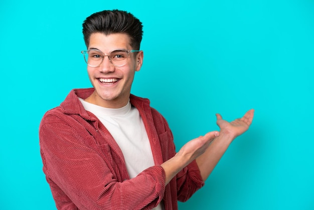 Young handsome caucasian man isolated on blue bakcground With glasses and presenting something