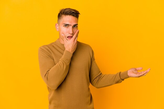 Young handsome caucasian man isolated holds copy space on a palm, keep hand over cheek. amazed and delighted