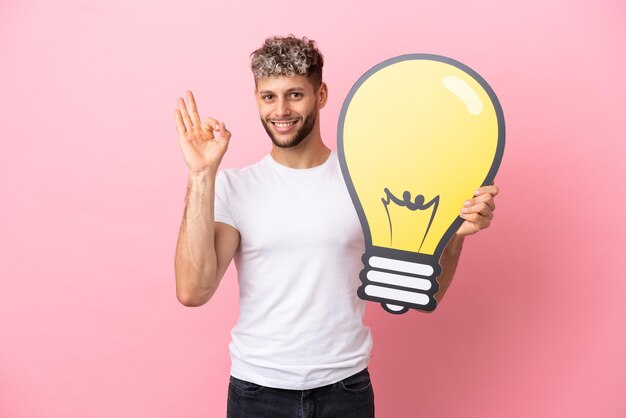 Young handsome caucasian man isolated on pink background holding a bulb icon and doing OK sign