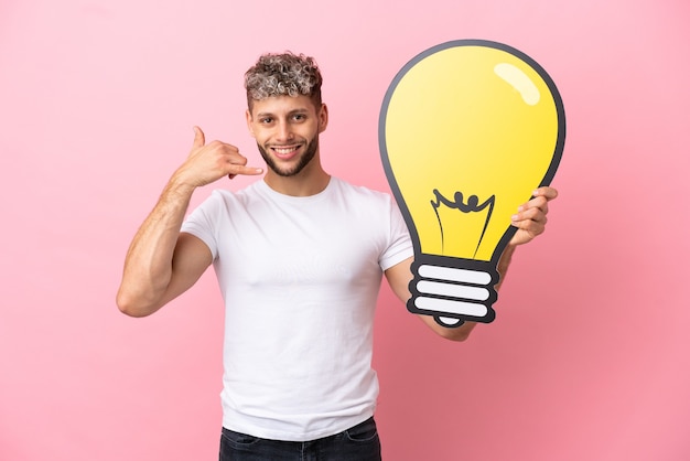 Young handsome caucasian man isolated on pink background holding a bulb icon and doing phone gesture