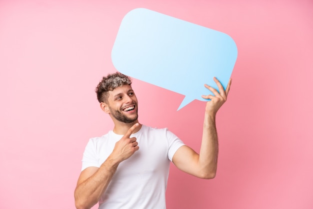 Young handsome caucasian man isolated on pink background holding an empty speech bubble and pointing it