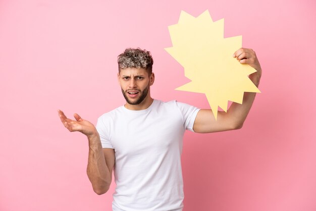 Young handsome caucasian man isolated on pink background holding an empty speech bubble and with frustrated expression