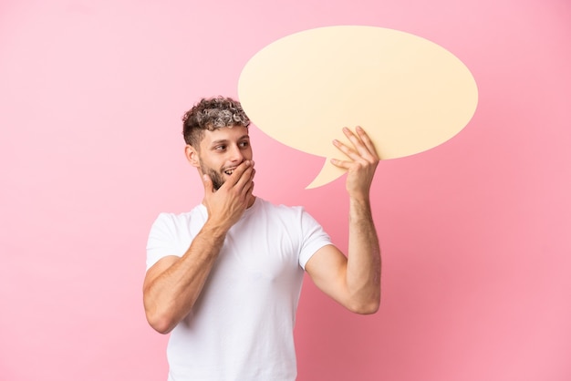 Young handsome caucasian man isolated on pink background holding an empty speech bubble with surprised expression