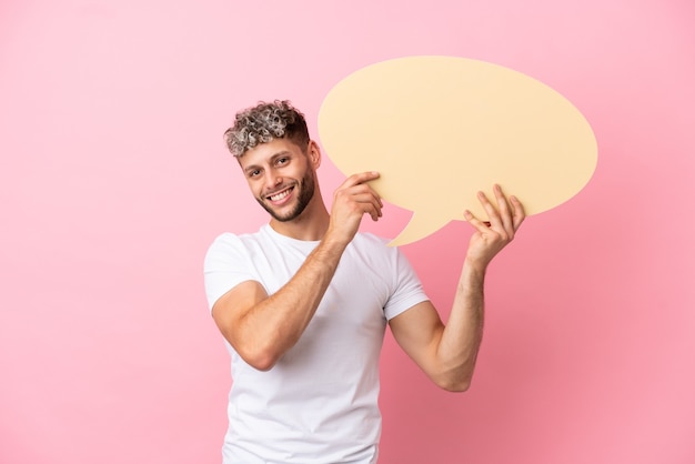 Young handsome caucasian man isolated on pink background holding an empty speech bubble