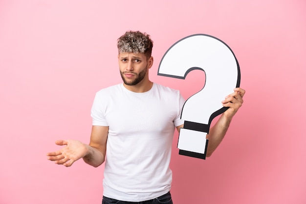 Young handsome caucasian man isolated on pink background holding a question mark icon and having doubts