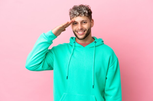 Young handsome caucasian man isolated on pink background saluting with hand with happy expression