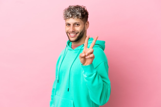 Young handsome caucasian man isolated on pink background smiling and showing victory sign