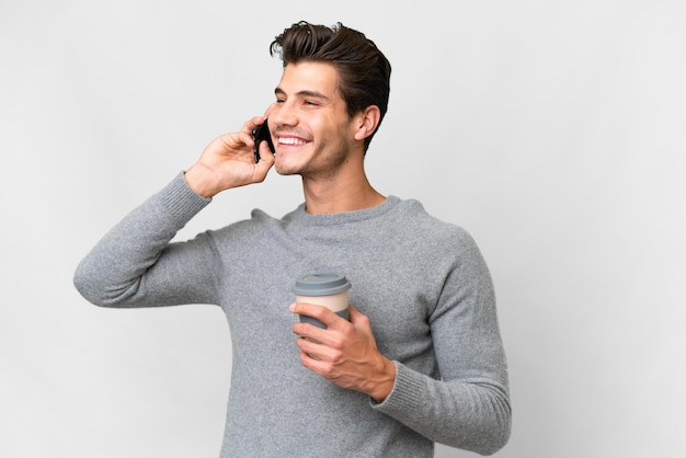 Молодой красивый кавказский мужчина на изолированном белом фоне держит кофе на вынос и мобильный телефон