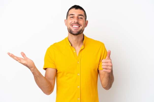 Young handsome caucasian man isolated on white background holding copyspace imaginary on the palm to insert an ad and with thumbs up