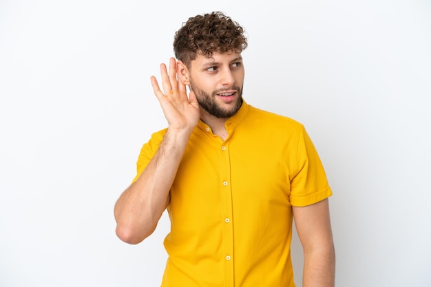 Foto giovane uomo caucasico bello isolato su sfondo bianco ascoltando qualcosa mettendo la mano sull'orecchio