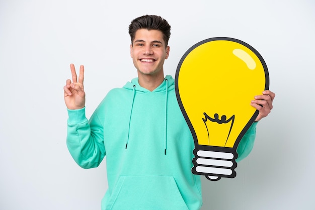 Young handsome caucasian man isolated on white bakcground holding a bulb icon and celebrating a victory