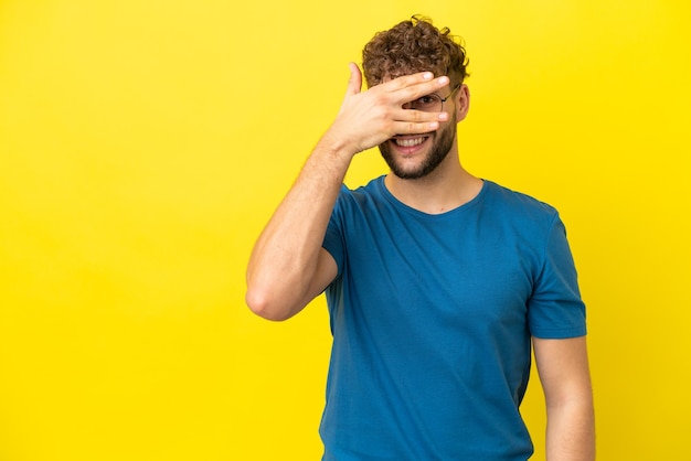 Young handsome caucasian man isolated on yellow background covering eyes by hands and smiling