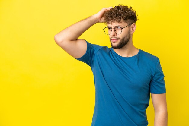 Young handsome caucasian man isolated on yellow background having doubts while scratching head