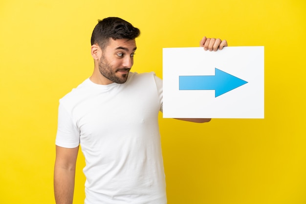 Young handsome caucasian man isolated on yellow background holding a placard with arrow symbol