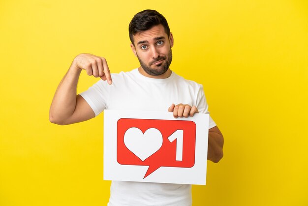 Young handsome caucasian man isolated on yellow background holding a placard with Like icon and pointing it