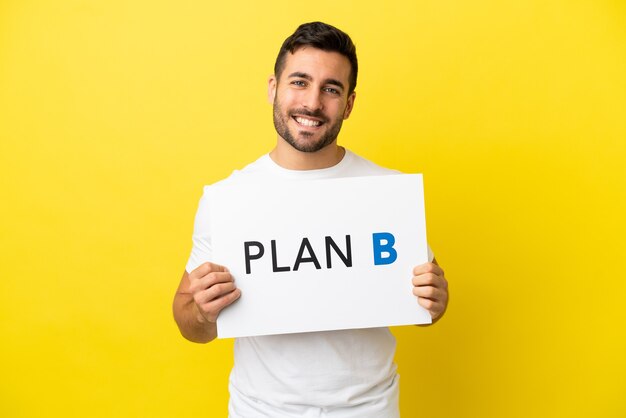 Young handsome caucasian man isolated on yellow background holding a placard with the message PLAN B with happy expression