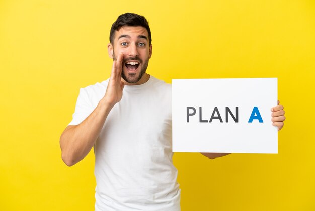 Young handsome caucasian man isolated on yellow background holding a placard with the message PLAN A and shouting