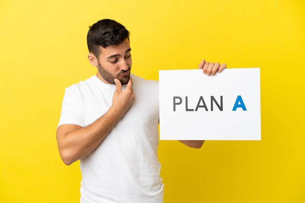 Young handsome caucasian man isolated on yellow background holding a placard with the message PLAN A and thinking