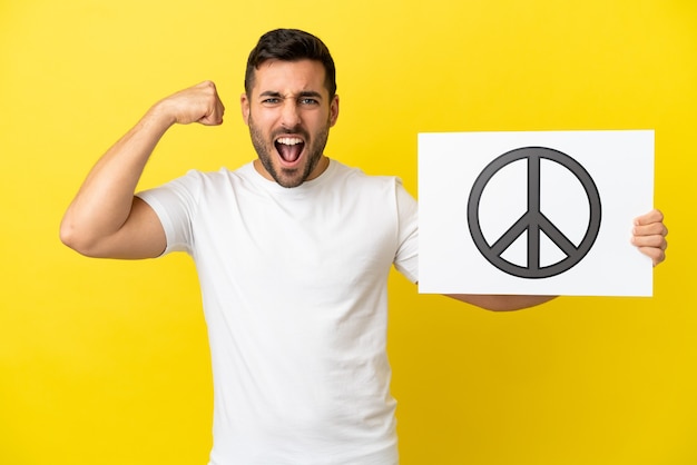 Young handsome caucasian man isolated on yellow background holding a placard with peace symbol and doing strong gesture