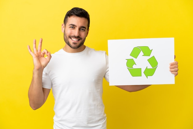 Young handsome caucasian man isolated on yellow background holding a placard with recycle icon and celebrating a victory