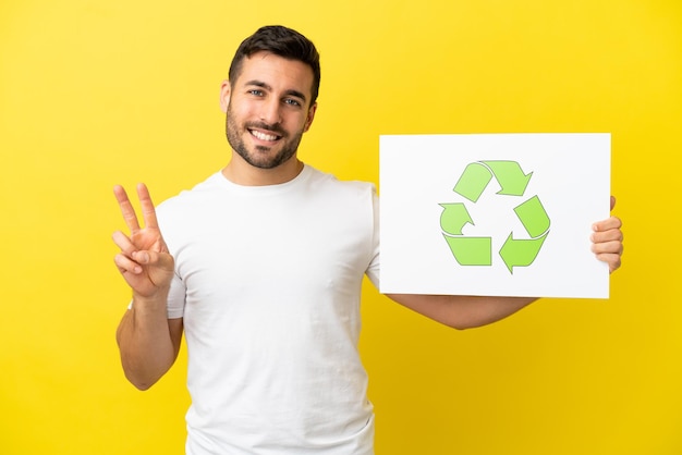 Young handsome caucasian man isolated on yellow background holding a placard with recycle icon and celebrating a victory