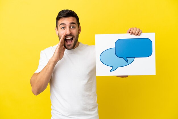 Young handsome caucasian man isolated on yellow background holding a placard with speech bubble icon and shouting