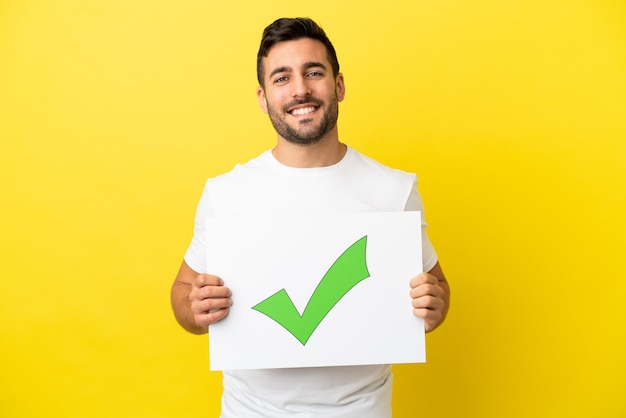 Young handsome caucasian man isolated on yellow background holding a placard with text Green check mark icon with happy expression