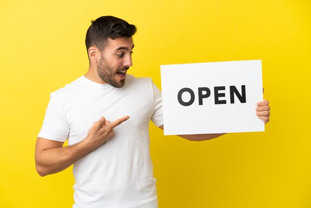 Young handsome caucasian man isolated on yellow background holding a placard with text OPEN and  pointing it