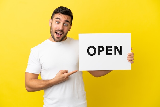 Young handsome caucasian man isolated on yellow background holding a placard with text OPEN and  pointing it