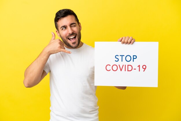 Young handsome caucasian man isolated on yellow background holding a placard with text Stop Covid 19 and doing phone gesture