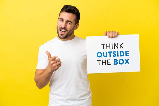 노란색 배경에 격리된 젊고 잘생긴 백인 남자는 Think Outside The Box라는 문구가 적힌 플래카드를 들고 앞을 가리키고 있습니다.