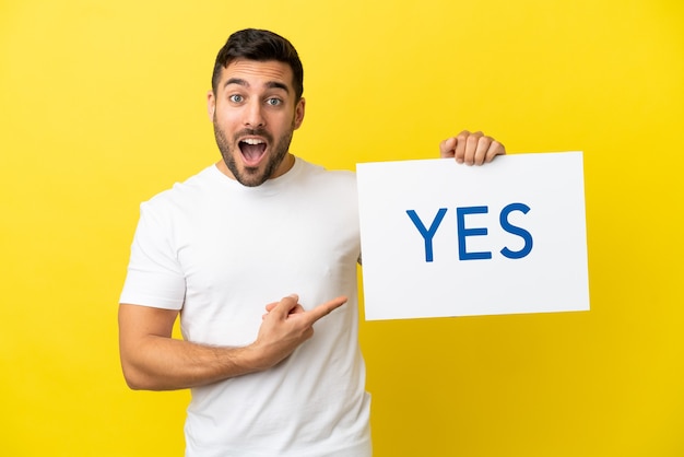 Young handsome caucasian man isolated on yellow background holding a placard with text YES and  pointing it