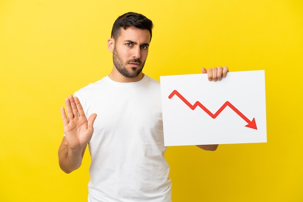 Young handsome caucasian man isolated on yellow background holding a sign with a decreasing statistics arrow symbol and doing stop sign