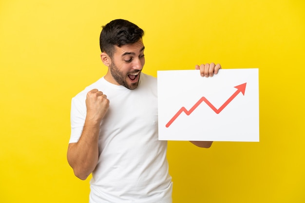 Young handsome caucasian man isolated on yellow background holding a sign with a growing statistics arrow symbol and celebrating a victory