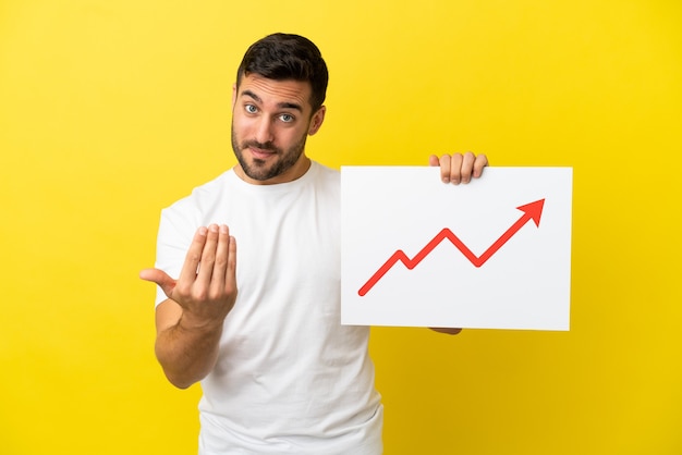 Young handsome caucasian man isolated on yellow background holding a sign with a growing statistics arrow symbol and doing coming gesture